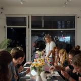 Group of people gathered around a table with flowers and food in an indoor setting.