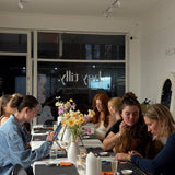 Group of people sitting around a table with various items on it, in a room with large windows.