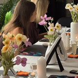 Two women at a table with floral arrangements and stationery items, in a room with plants and a recycling sign.