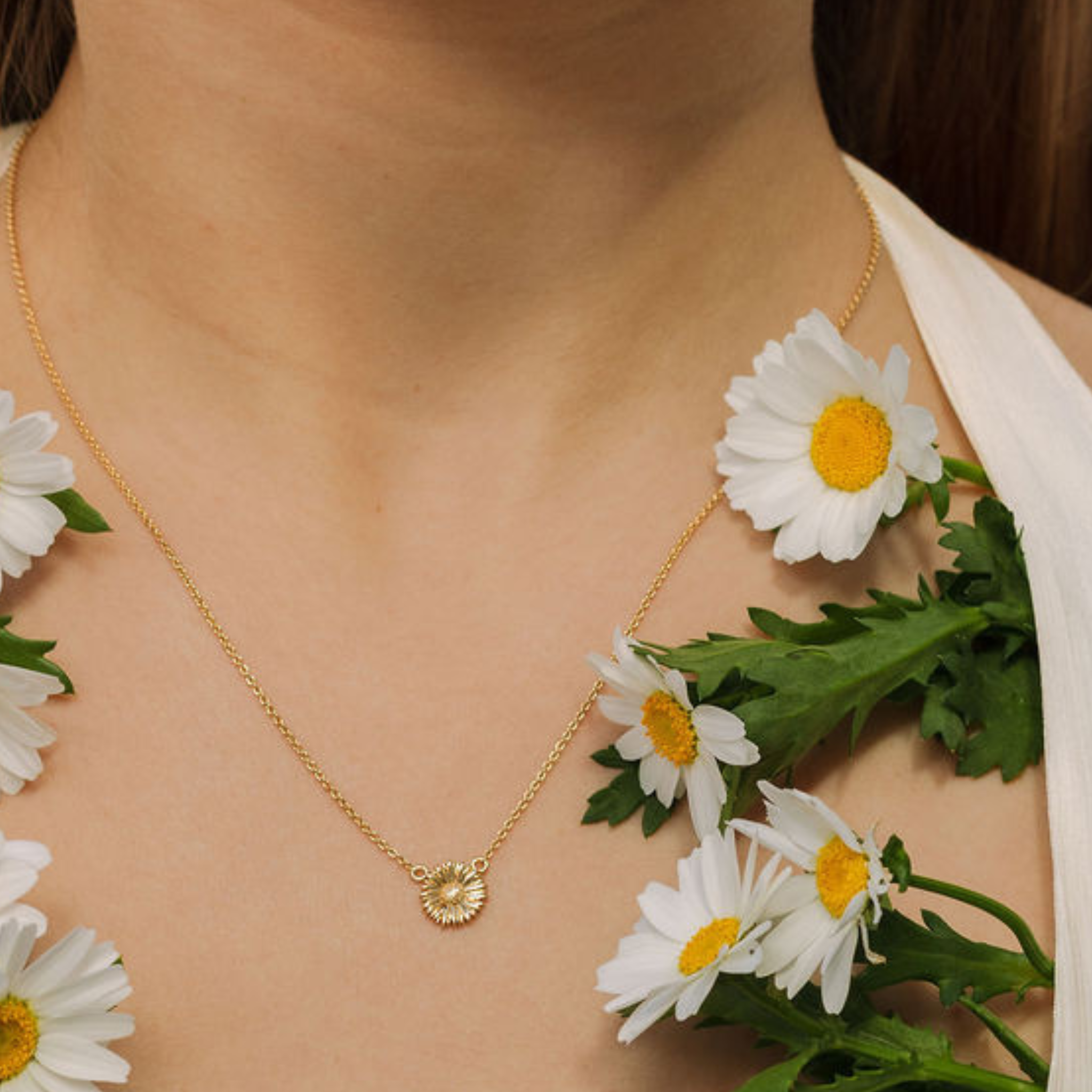 Gold necklace with a daisy pendant worn by a person surrounded by daisies.