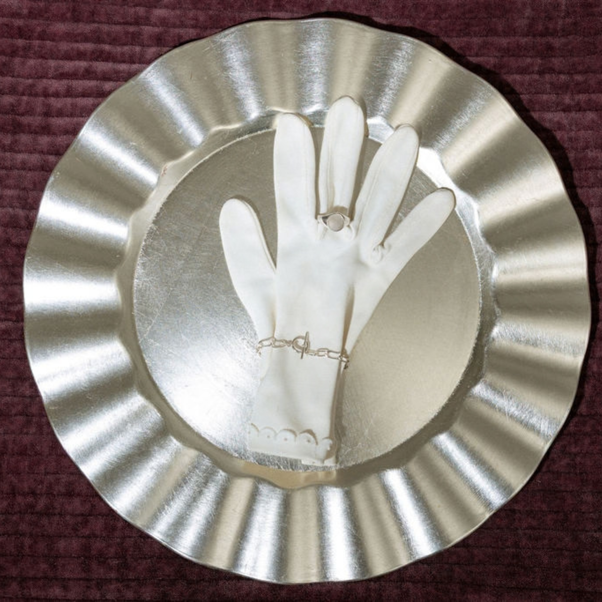 A white glove displayed on a metallic silver stand, with a delicate silver ring on the glove's wrist and a small silver charm on the back of the hand, set against a dark burgundy textured background.