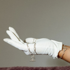 A white glove-covered hand holds a delicate silver chain bracelet with a toggle clasp against a plain taupe background.