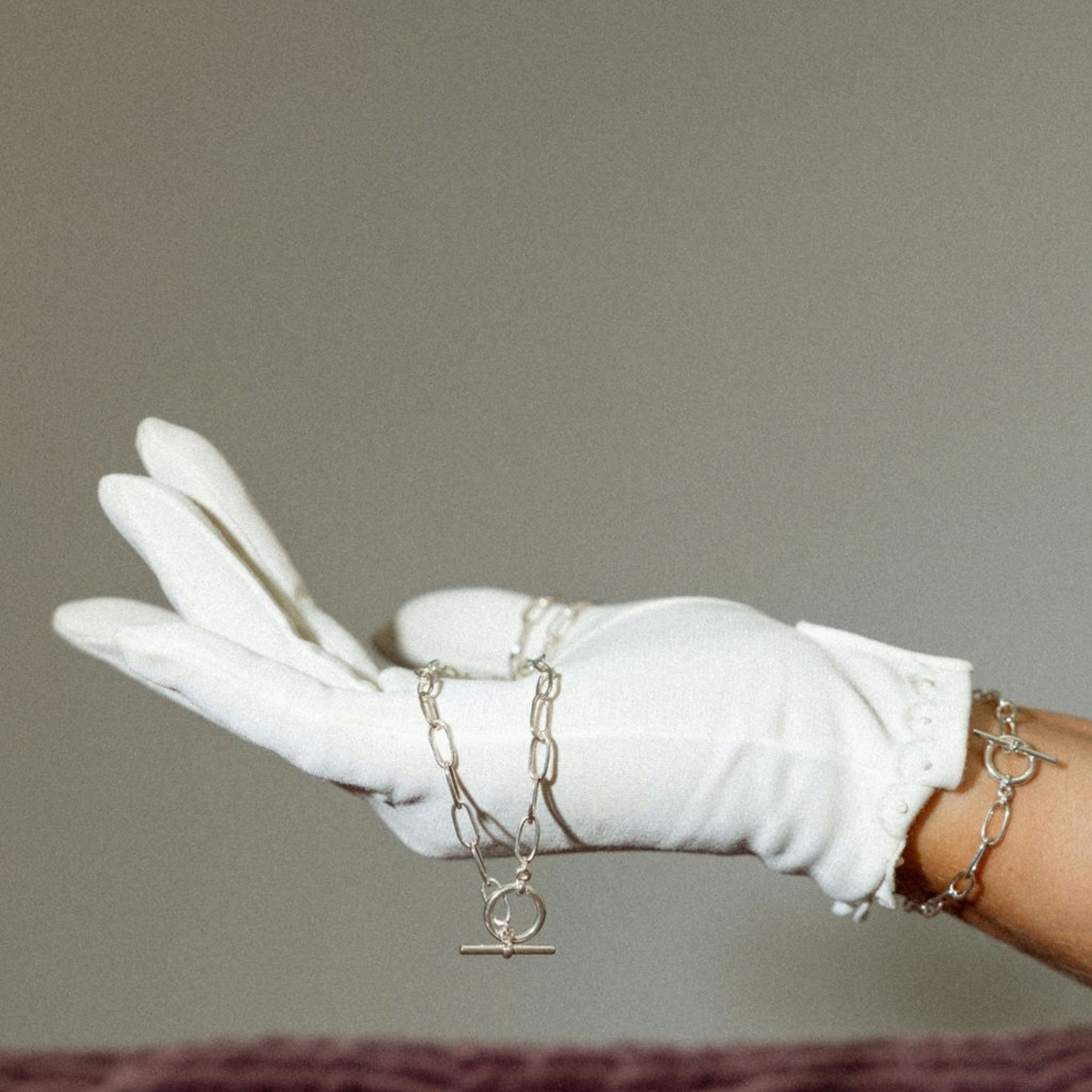 A white glove-covered hand holds a delicate silver chain bracelet with a toggle clasp against a plain taupe background.