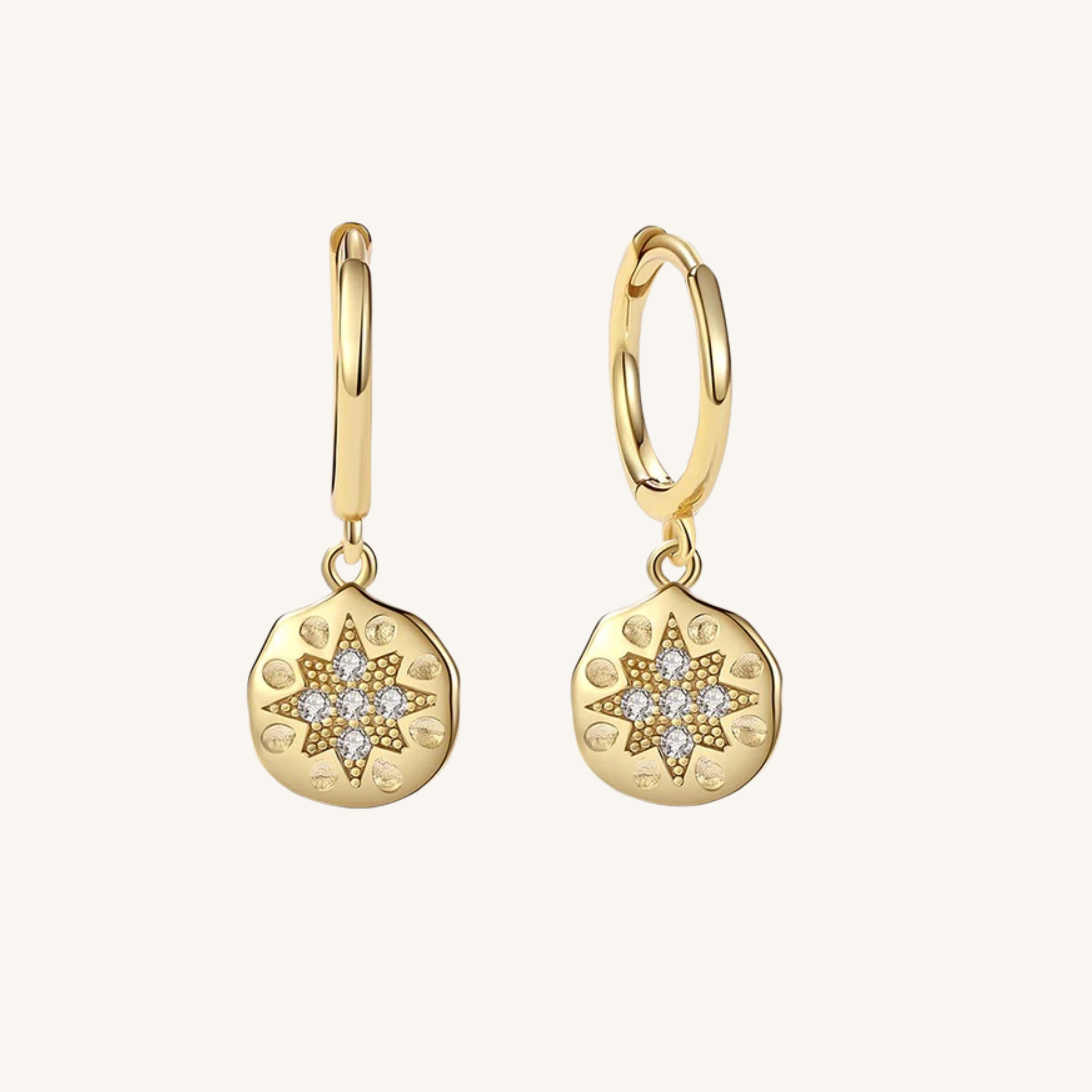 A pair of gold huggie hoop earrings with round, textured gold pendants featuring a starburst design paved with small clear stones, set against a solid off-white background.
