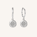 A pair of silver huggie hoop earrings with circular starburst charms, set against a solid off-white background.