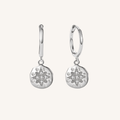 A pair of silver huggie hoop earrings with circular starburst charms, set against a solid off-white background.