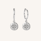 A pair of silver huggie hoop earrings with circular starburst charms, set against a solid off-white background.