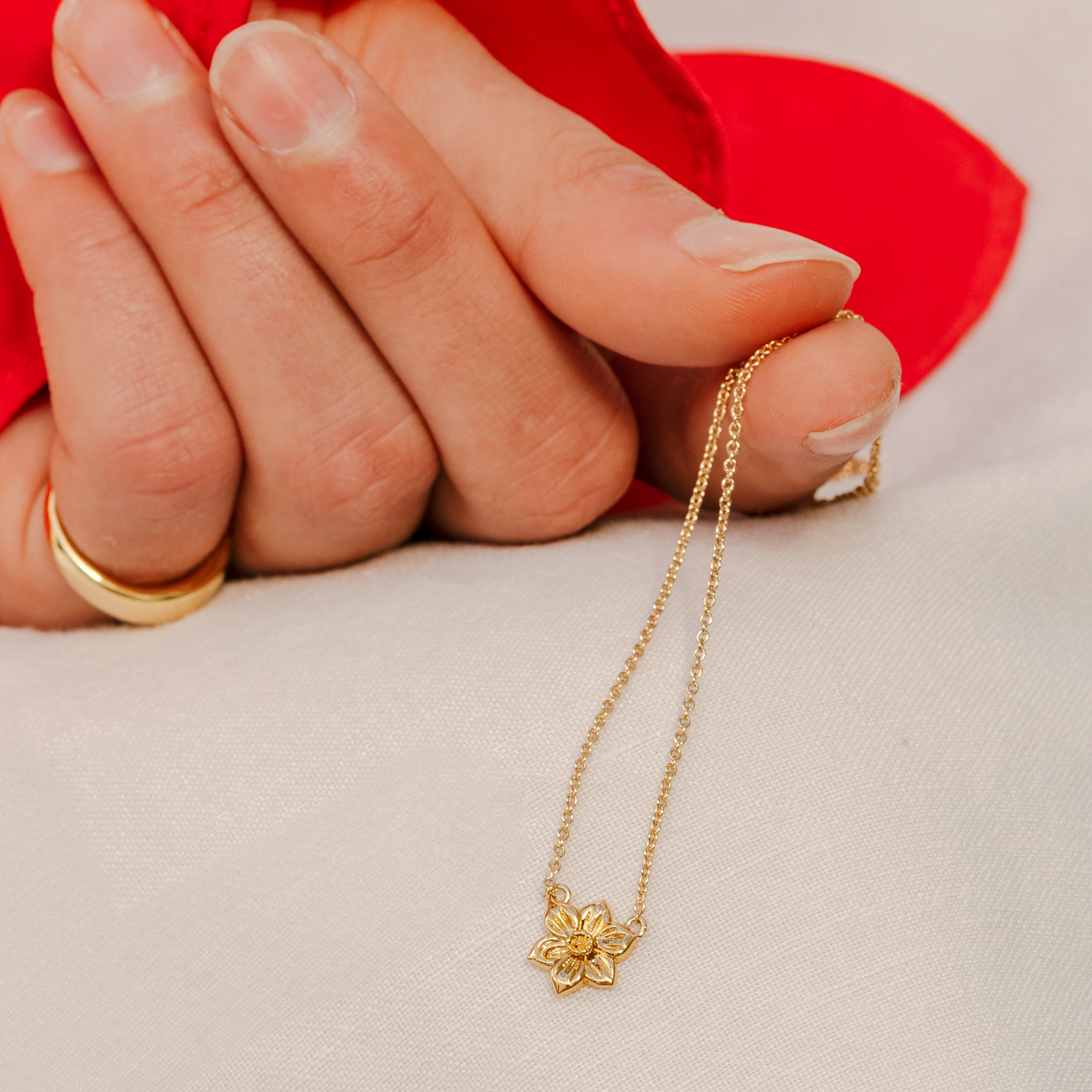 Gold necklace with a flower pendant held by a hand against a red fabric background