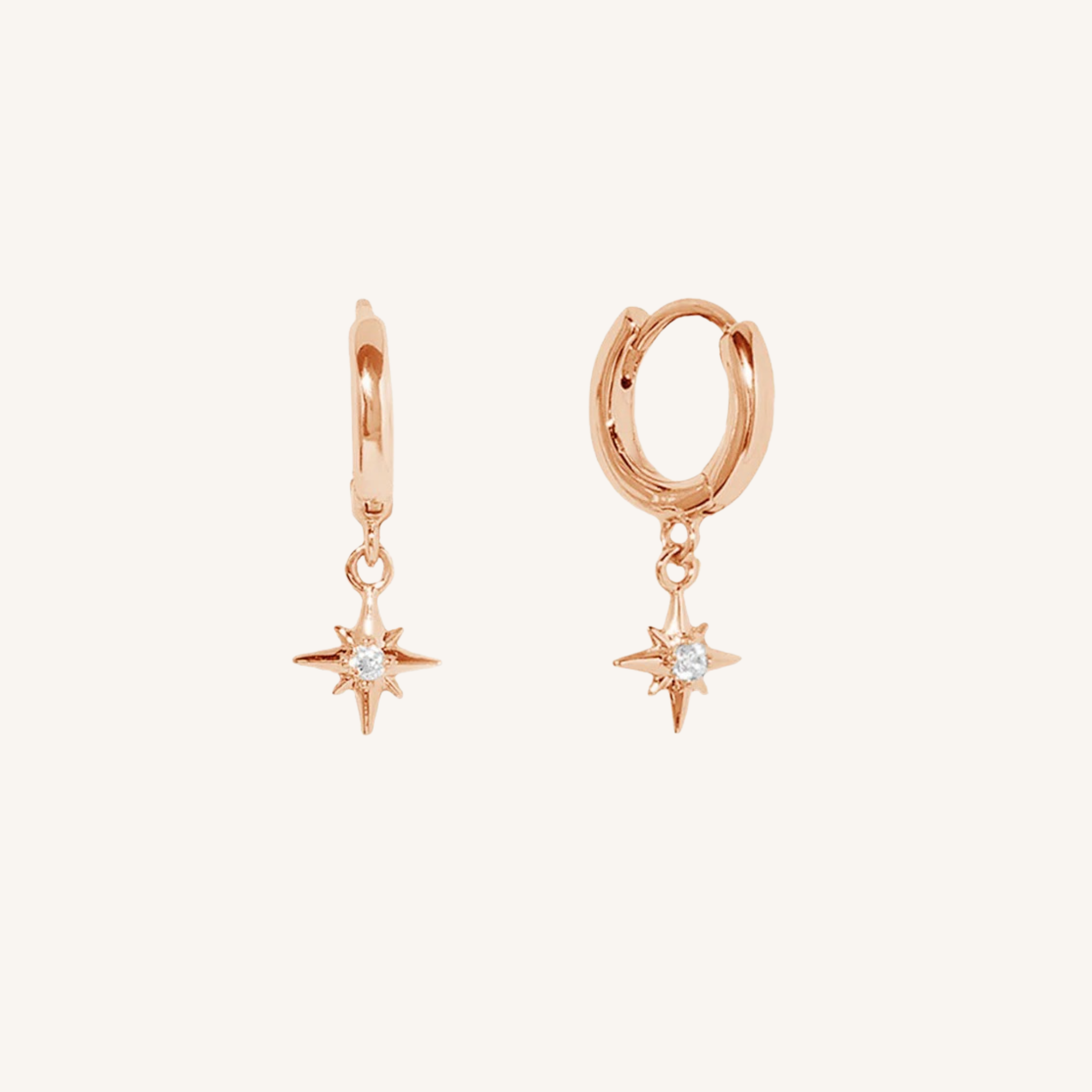 A pair of rose gold huggie hoop earrings with dangling six-pointed star charms set with small clear gemstones, shown on a light cream background.