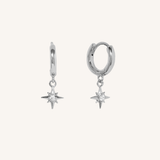 A pair of silver huggie hoop earrings with dangling starburst charms, shown on a plain off-white background.