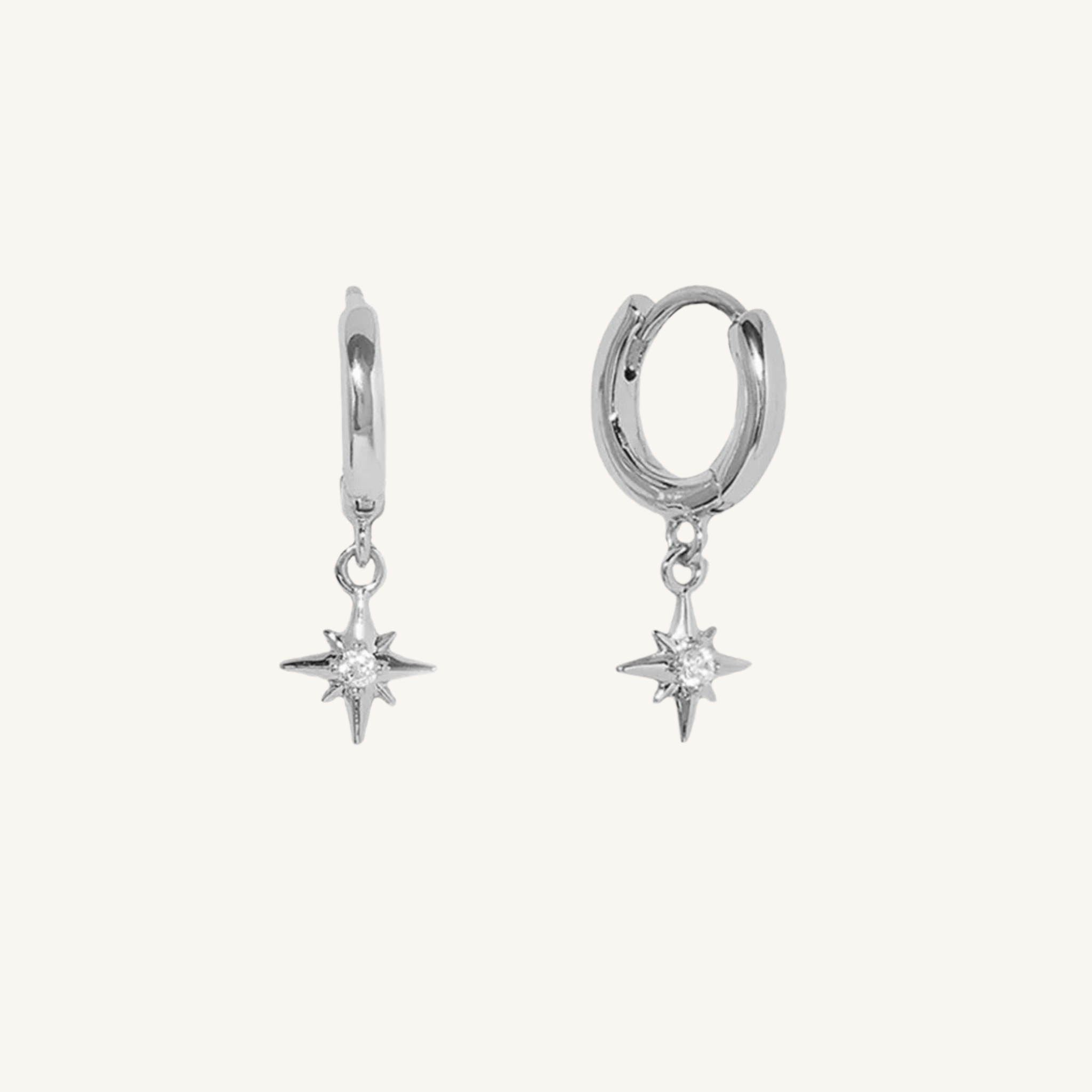 A pair of silver huggie hoop earrings with dangling starburst charms, shown on a plain off-white background.