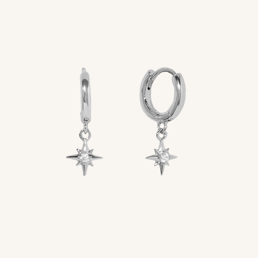 A pair of silver huggie hoop earrings with dangling starburst charms, shown on a plain off-white background.
