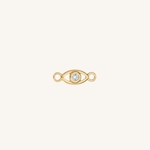 A gold eye-shaped charm with a clear round gemstone in the center, set against a solid off-white background.