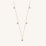A delicate gold chain necklace featuring five small, round clear gemstones spaced along the chain, displayed on a light cream background.