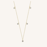 A delicate gold chain necklace featuring five small, round clear gemstones spaced along the chain, displayed on a light cream background.