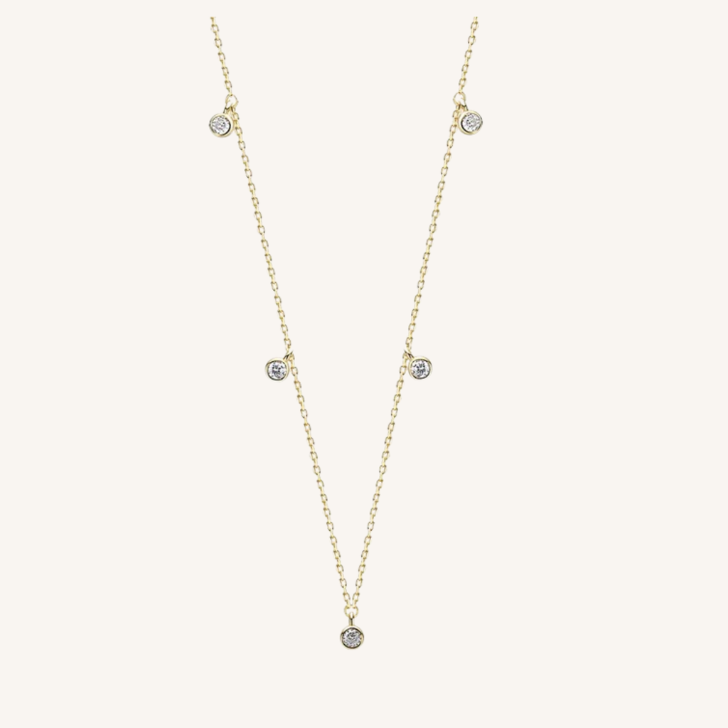 A delicate gold chain necklace featuring five small, round clear gemstones spaced along the chain, displayed on a light cream background.