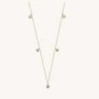 A delicate gold chain necklace featuring five small, round clear gemstones spaced along the chain, displayed on a light cream background.