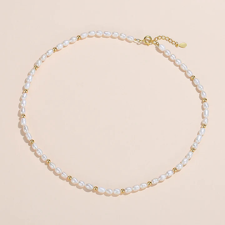 A delicate pearl necklace with small gold spacer beads and a gold clasp, displayed on a light cream background.