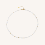 A delicate pearl necklace with small gold spacer beads and a gold clasp, displayed on a light cream background.