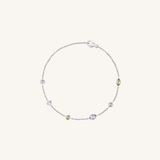 A delicate silver chain bracelet with five small, colorful gemstone accents in purple, green, and clear, displayed on a light cream background.