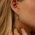 A close-up of a woman's ear and neck wearing a delicate gold huggie hoop earring with a small horizontal bar and a tiny clear gemstone at the center.