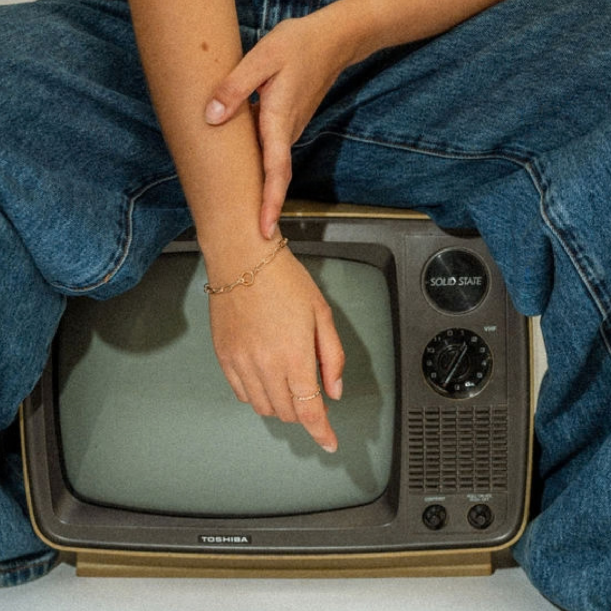 A person wearing blue jeans sits behind a vintage Toshiba television, with one arm resting across the TV. A delicate gold chain bracelet is visible on the wrist.