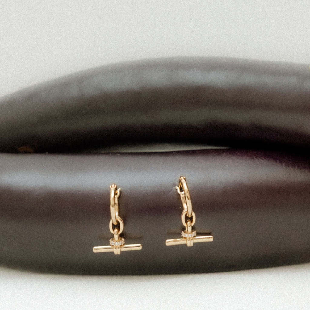 A pair of small gold huggie hoop earrings with a barbell charm, displayed on a dark brown textured surface against a light gray background.