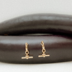 A pair of small gold huggie hoop earrings with a barbell charm, displayed on a dark brown textured surface against a light gray background.