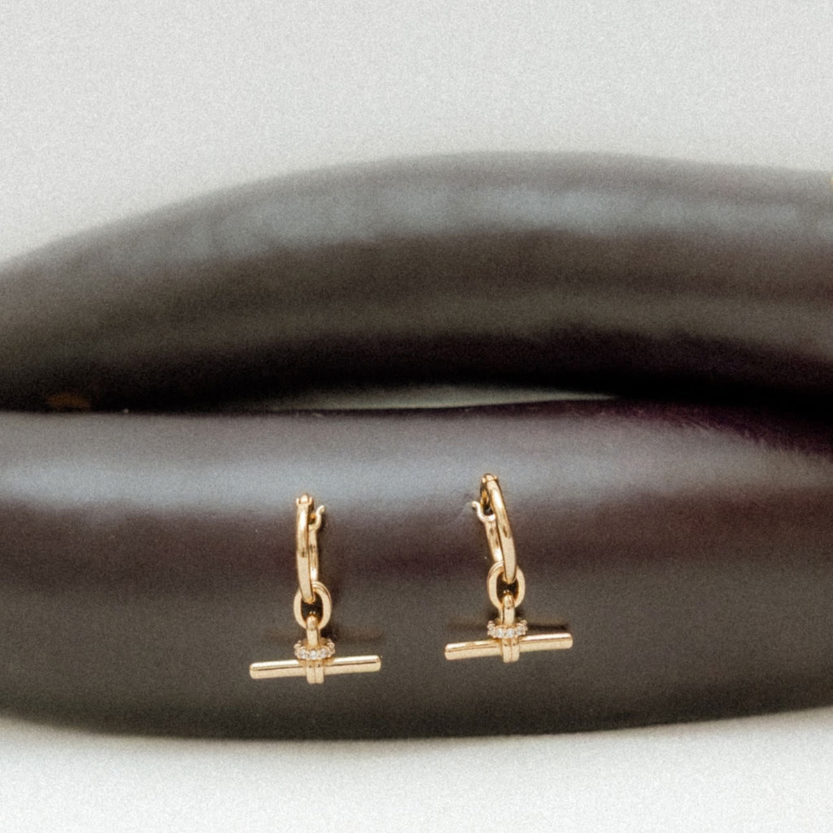 A pair of small gold huggie hoop earrings with a barbell charm, displayed on a dark brown textured surface against a light gray background.