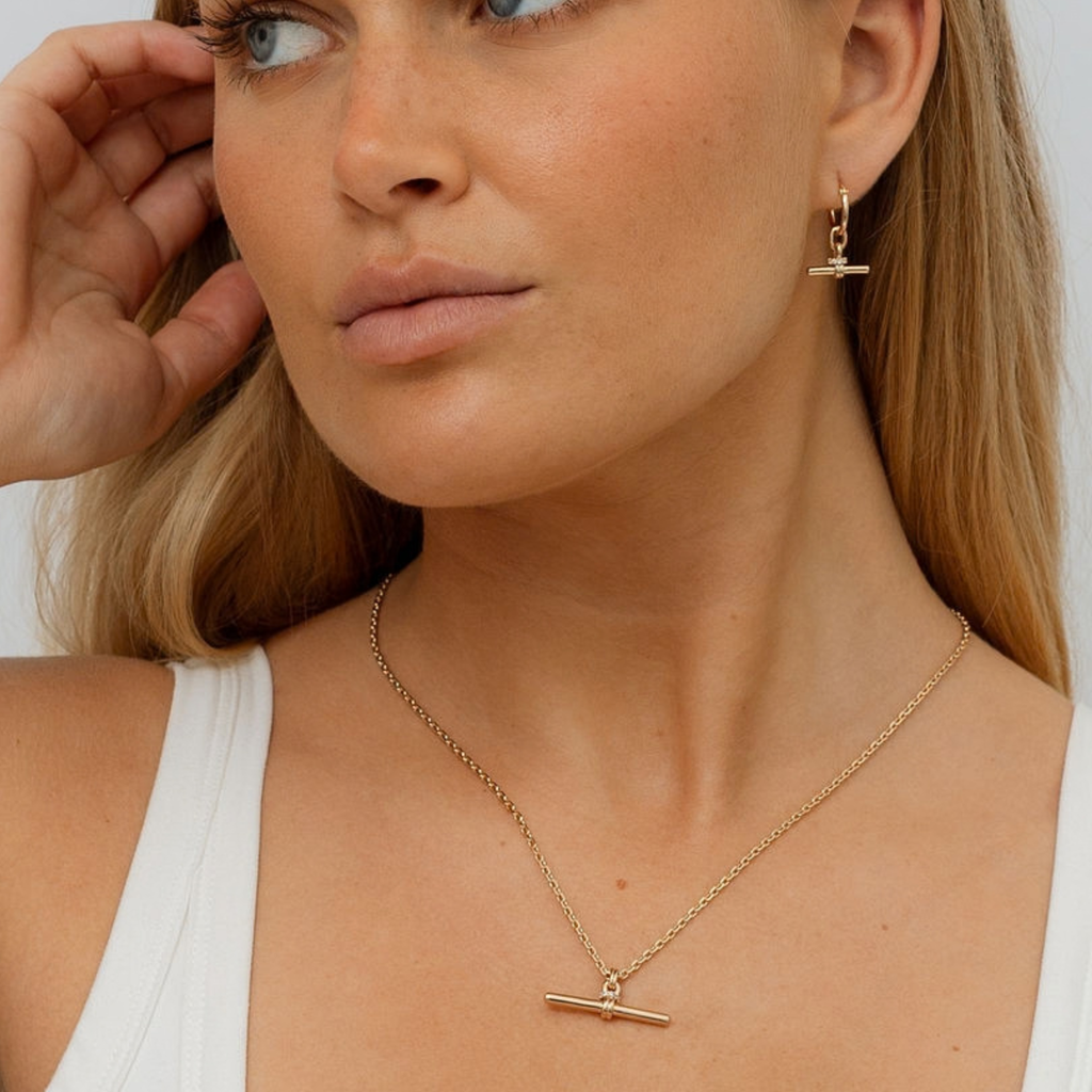 A close-up lifestyle shot of a woman wearing a delicate gold necklace and matching drop earrings with horizontal bar pendants, styled with a white tank top.