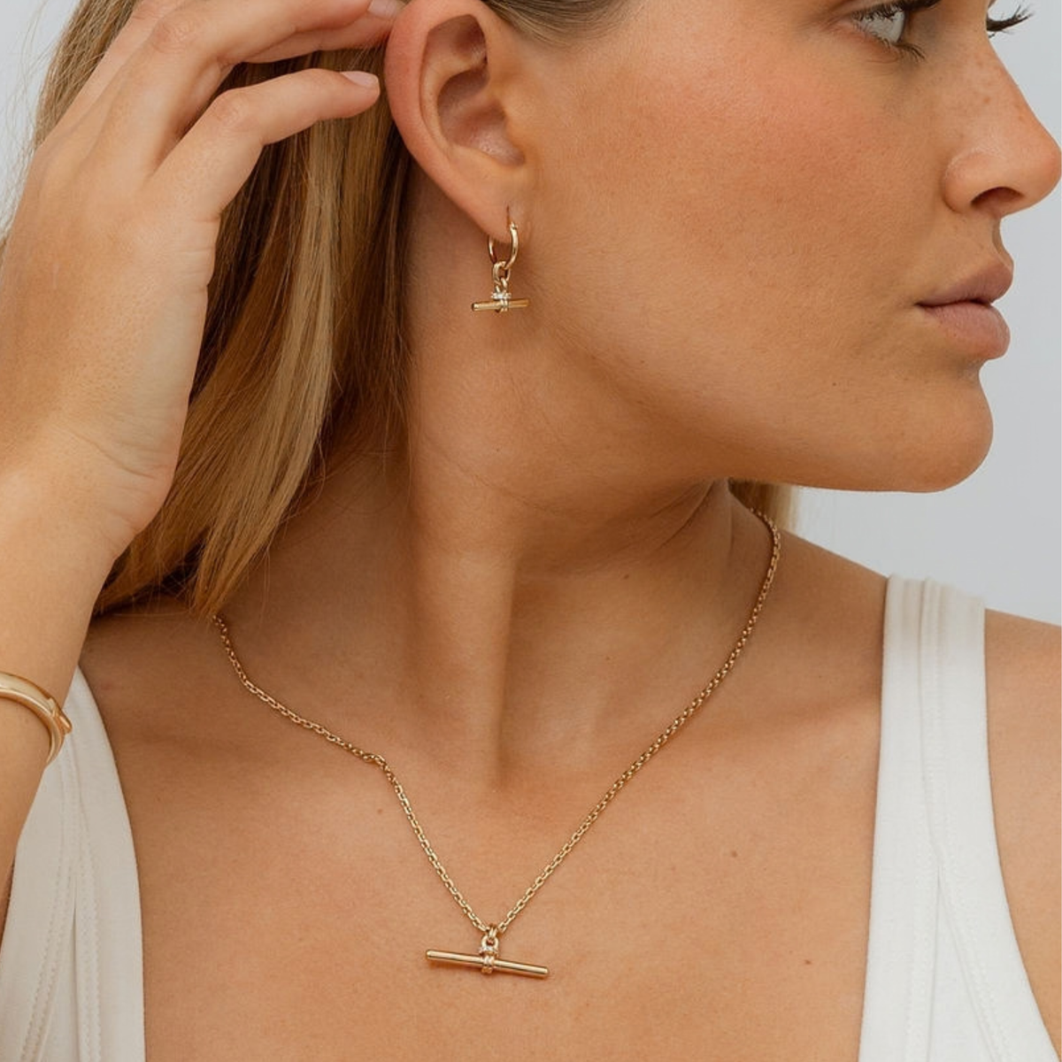 A close-up lifestyle shot of a model wearing a delicate gold necklace and matching small drop earrings with horizontal bar charms, styled with a white tank top.