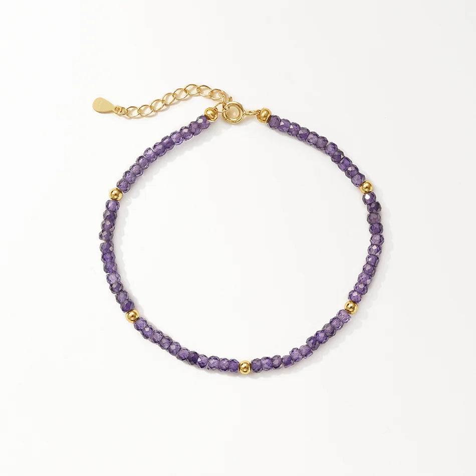 A delicate beaded bracelet featuring faceted purple beads and small gold spacer beads, with a gold clasp and extender chain, displayed on a light cream background.