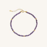 A delicate beaded bracelet featuring faceted purple beads and small gold spacer beads, with a gold clasp and extender chain, displayed on a light cream background.