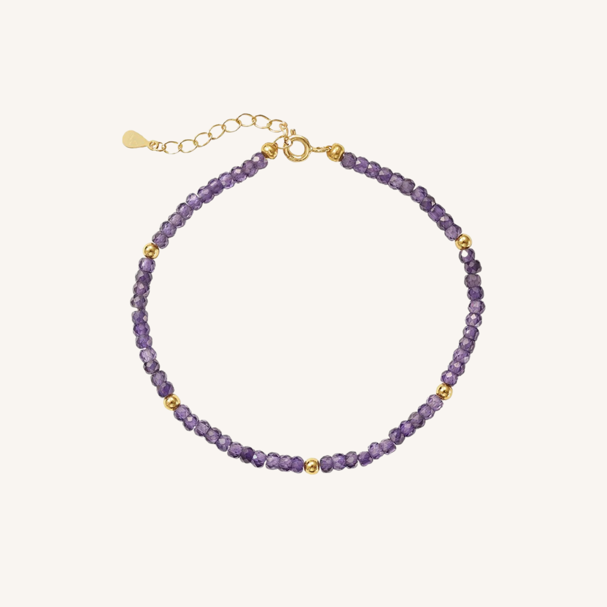 A delicate beaded bracelet featuring faceted purple beads and small gold spacer beads, with a gold clasp and extender chain, displayed on a light cream background.