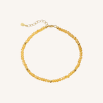 A delicate beaded necklace made of small, faceted translucent yellow beads with gold spacer beads and a gold clasp with an extender chain, shown on a light cream background.