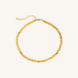 A delicate beaded necklace made of small, faceted translucent yellow beads with gold spacer beads and a gold clasp with an extender chain, shown on a light cream background.