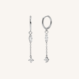 A pair of silver huggie hoop earrings with dangling chains and clear gemstone charms, displayed against a solid off-white background.