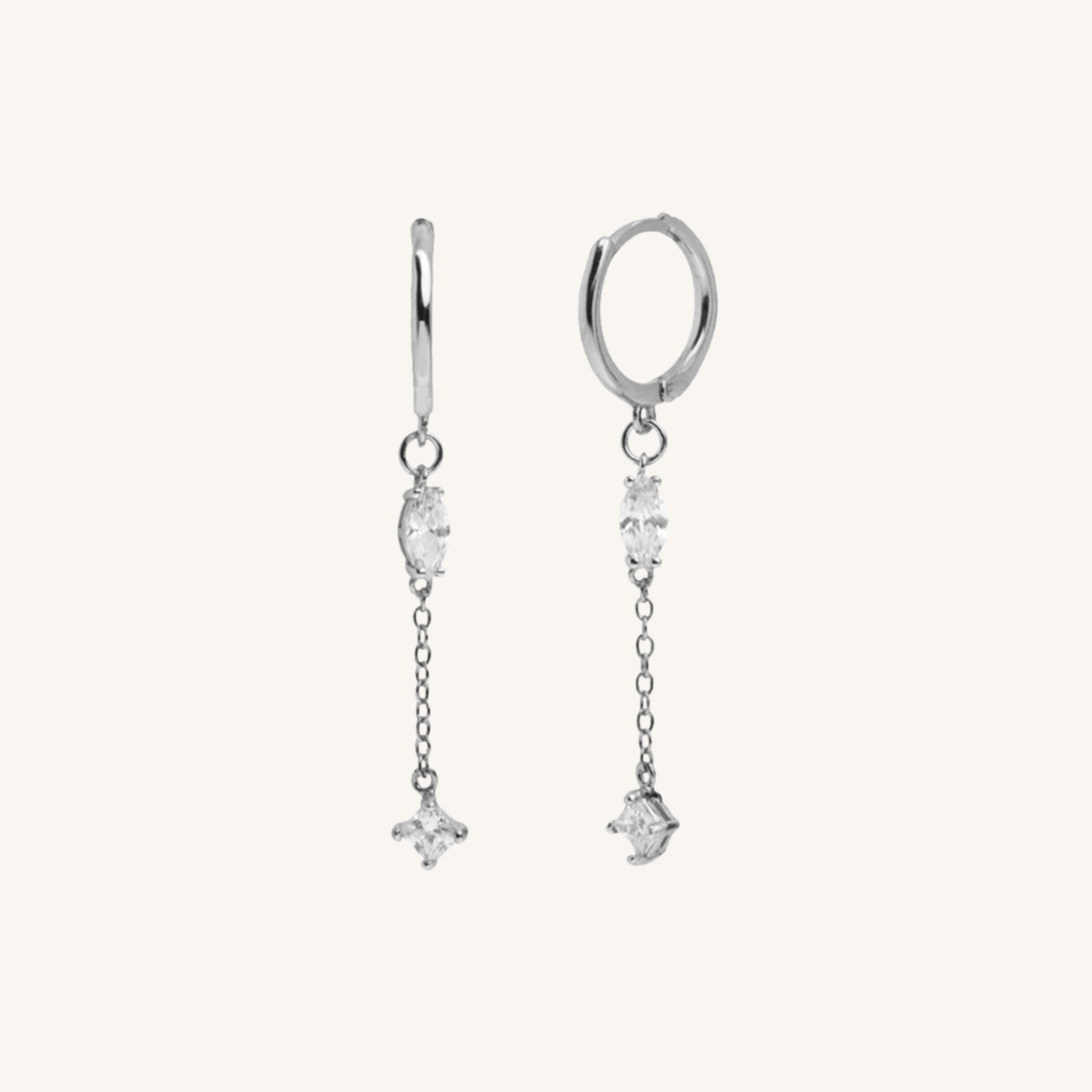 A pair of silver huggie hoop earrings with dangling chains and clear gemstone charms, displayed against a solid off-white background.