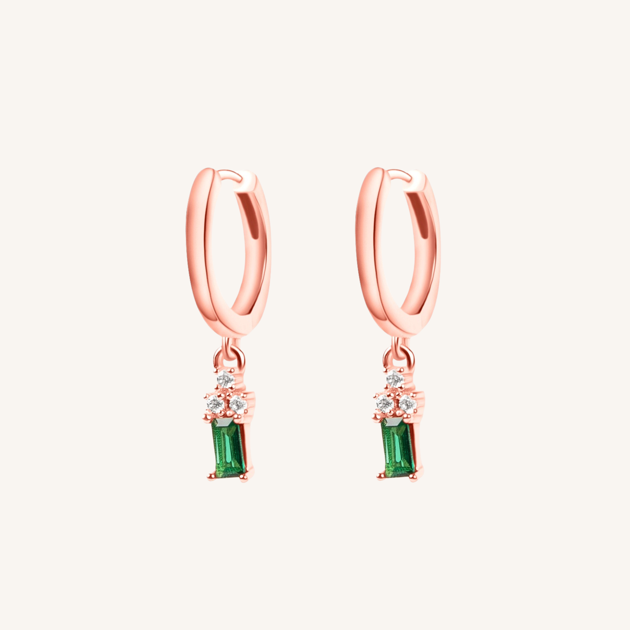 A pair of rose gold huggie hoop earrings with a dangling emerald green stone and small clear accent stones, shown on a light cream background.