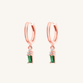 A pair of rose gold huggie hoop earrings with a dangling emerald green stone and small clear accent stones, shown on a light cream background.