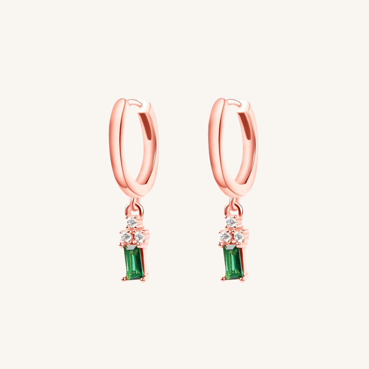 A pair of rose gold huggie hoop earrings with a dangling emerald green stone and small clear accent stones, shown on a light cream background.