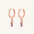 A pair of rose gold hoop earrings with a dangling purple amethyst and small clear cubic zirconia accents, shown on a light cream background.