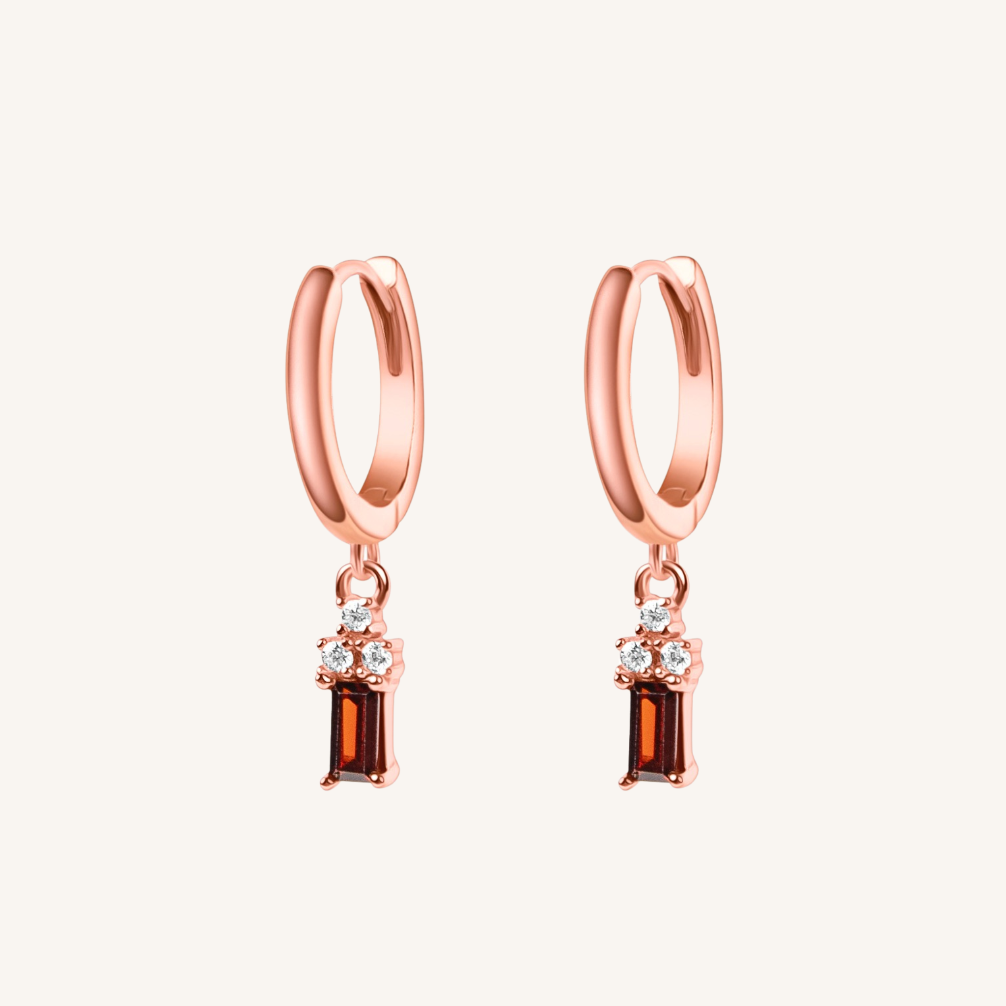 A pair of rose gold huggie hoop earrings with small clear crystal clusters and rectangular brown drops, shown on a light cream background.