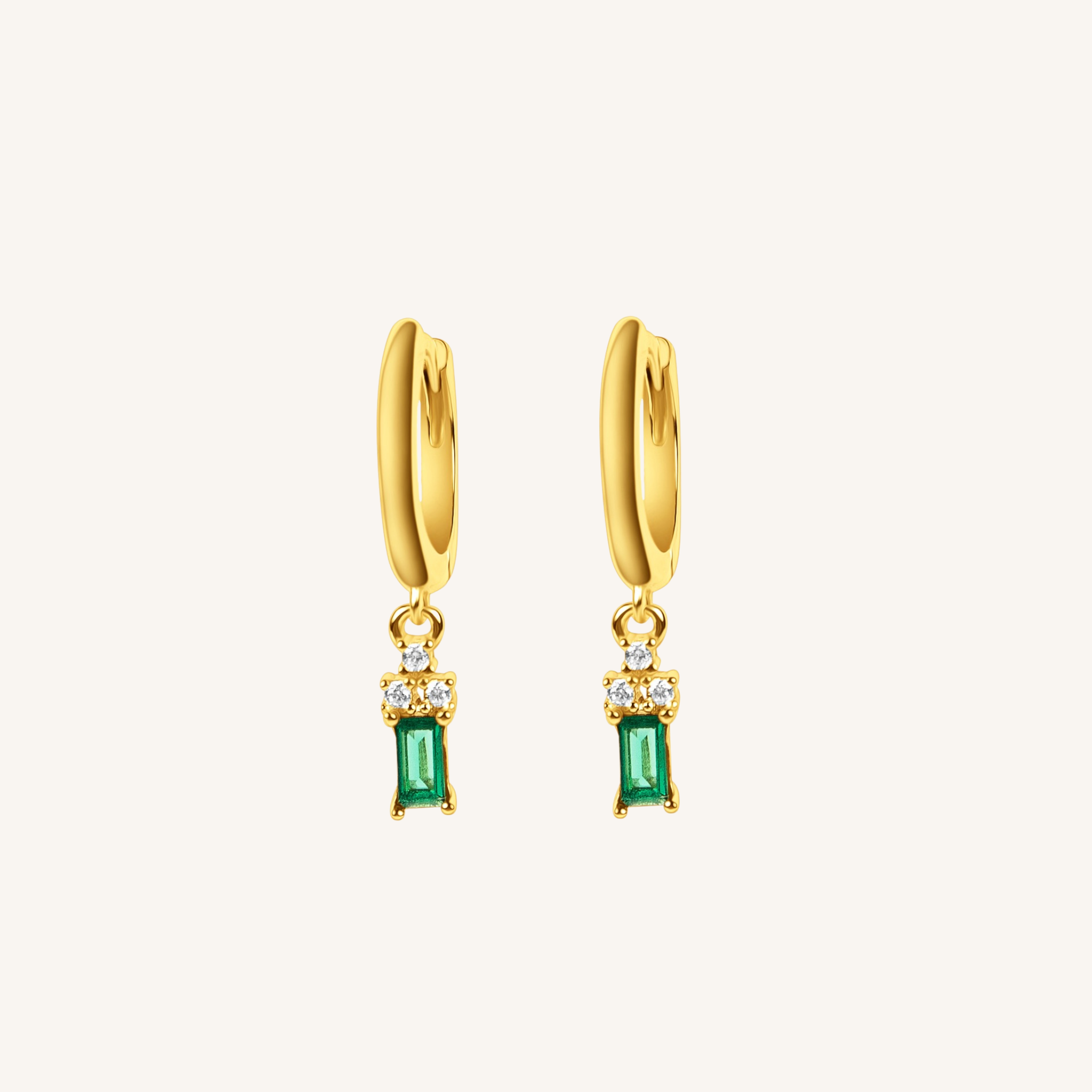 A pair of gold huggie hoop earrings with a dangling emerald-cut green stone and small clear accent stones, shown on a light cream background.