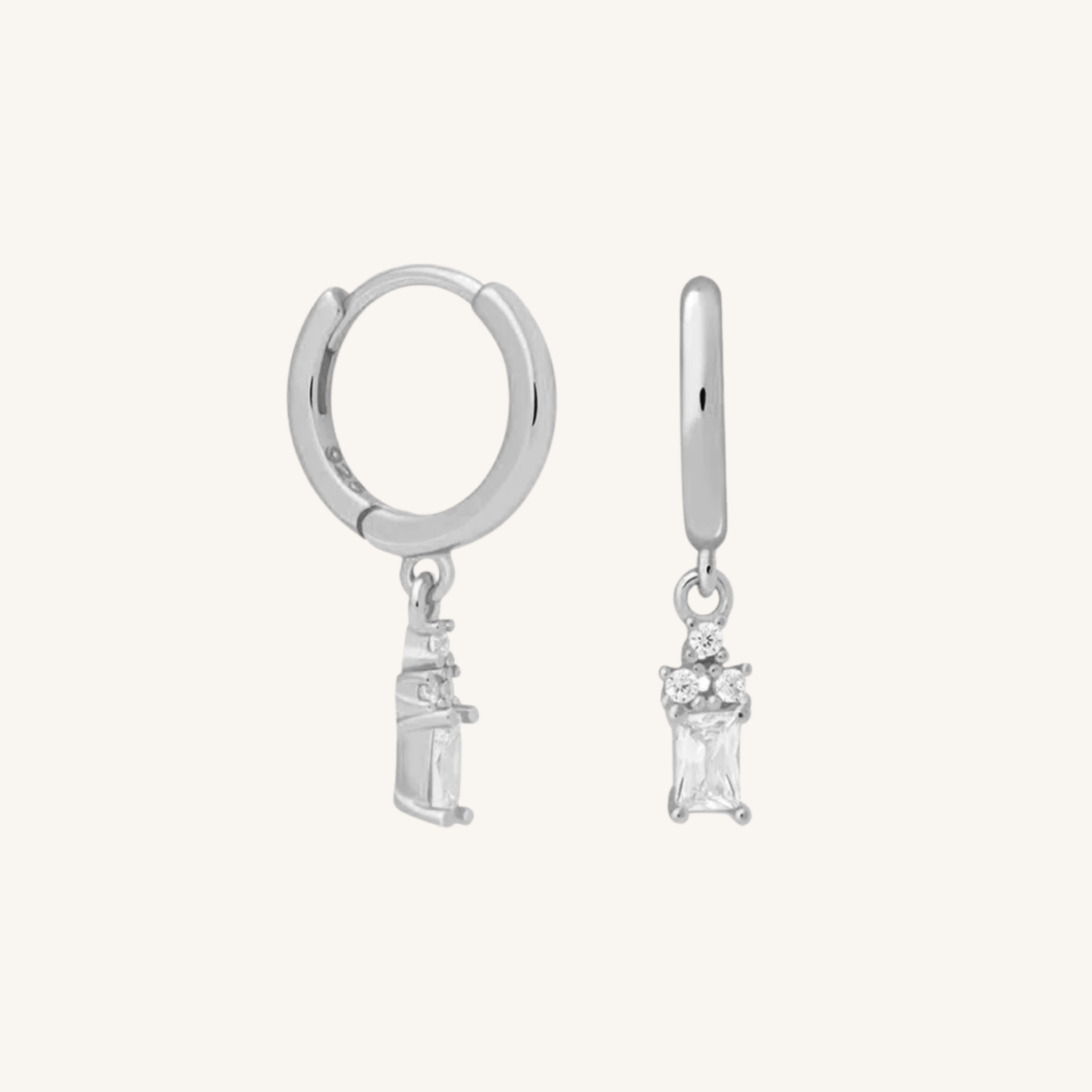 A pair of silver huggie hoop earrings with a dangling clear gemstone, shown on a light cream background.