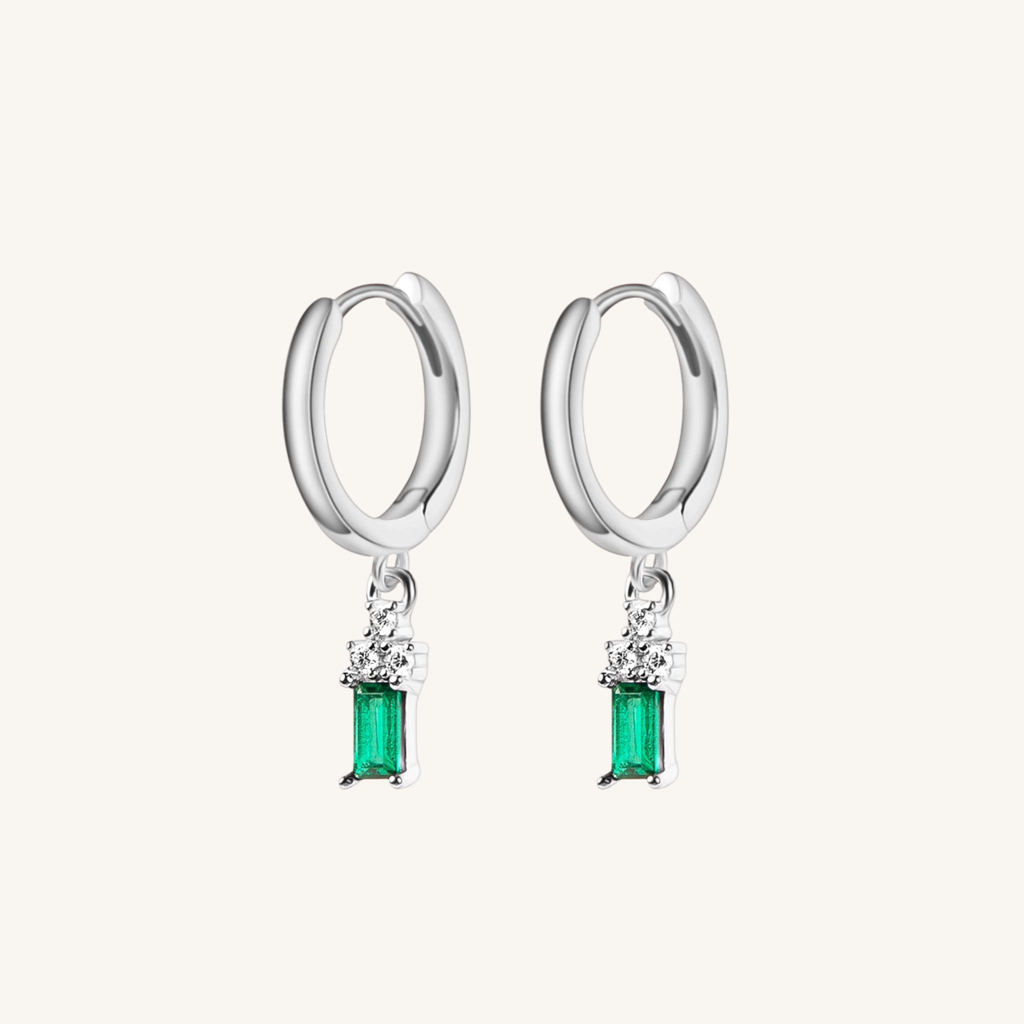 A pair of silver huggie hoop earrings with a dangling emerald-cut green gemstone and small clear accent stones, shown on a light cream background.
