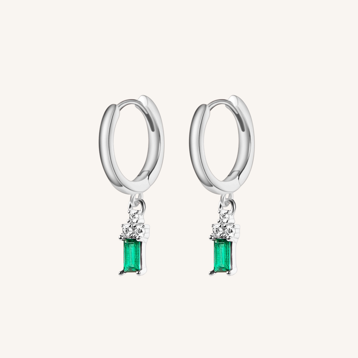 A pair of silver huggie hoop earrings with a dangling emerald-cut green gemstone and small clear accent stones, shown on a light cream background.