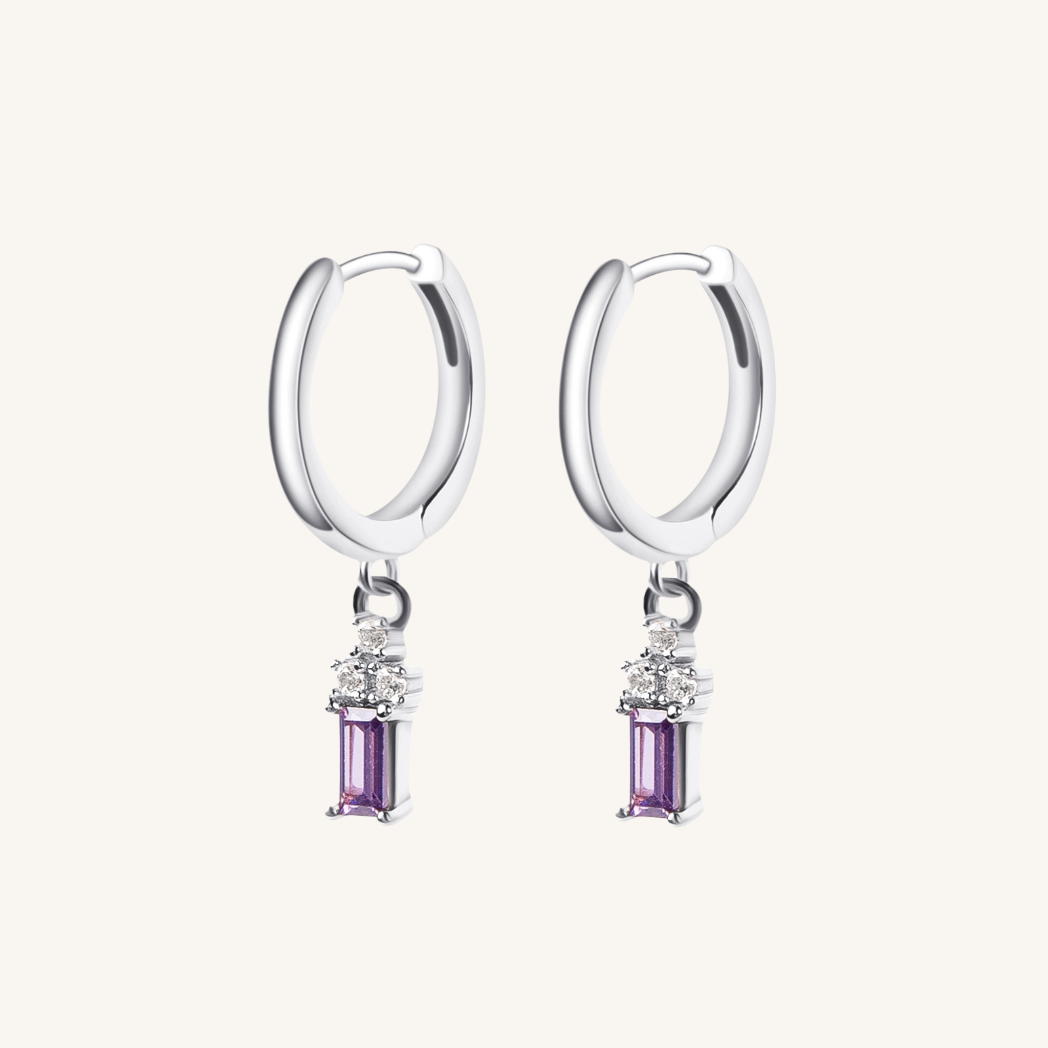 A pair of silver huggie hoop earrings with a dangling rectangular purple gemstone and small clear accent stones, shown on a light cream background.