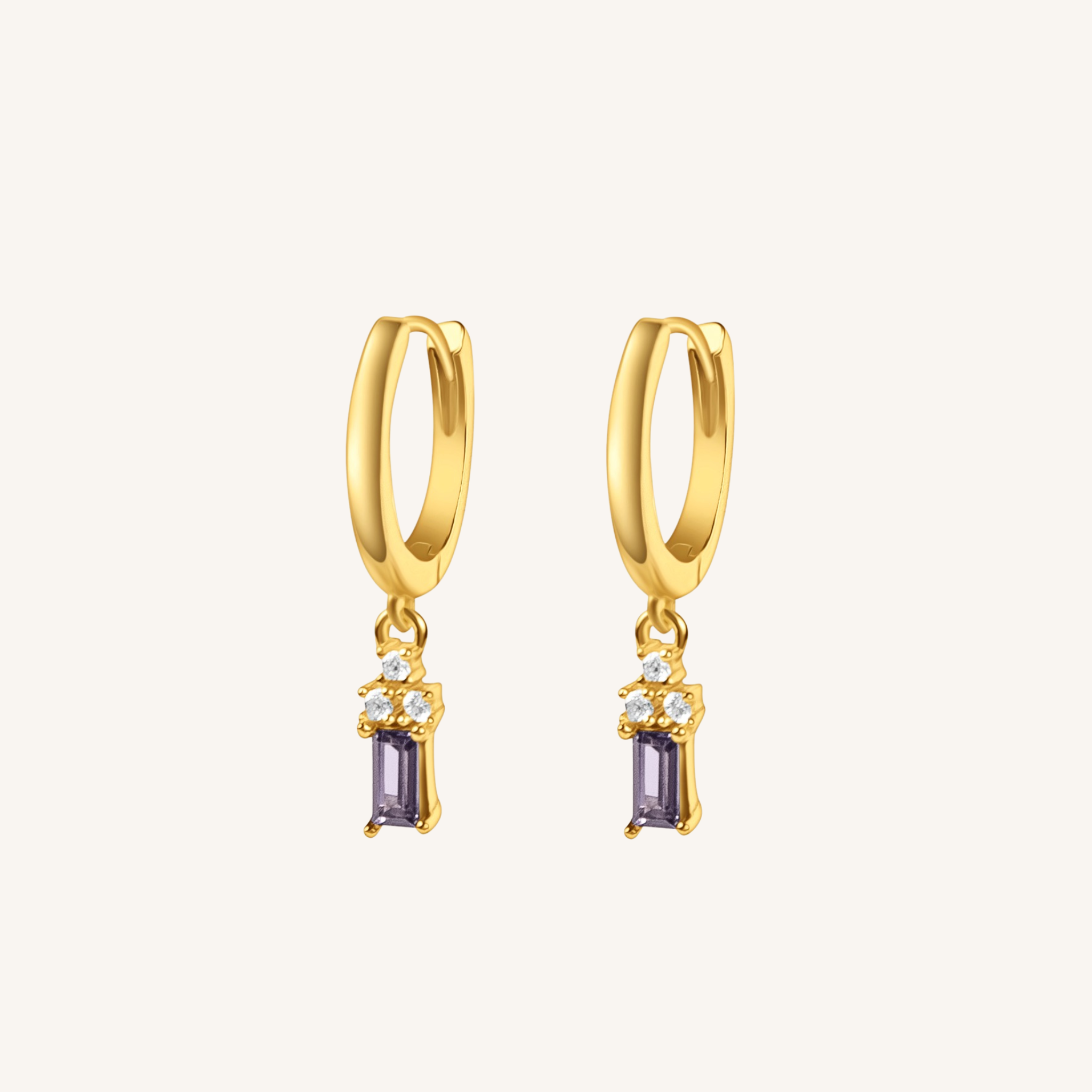 A pair of gold huggie hoop earrings with a dangling rectangular purple gemstone and small clear accent stones, shown on a light cream background.