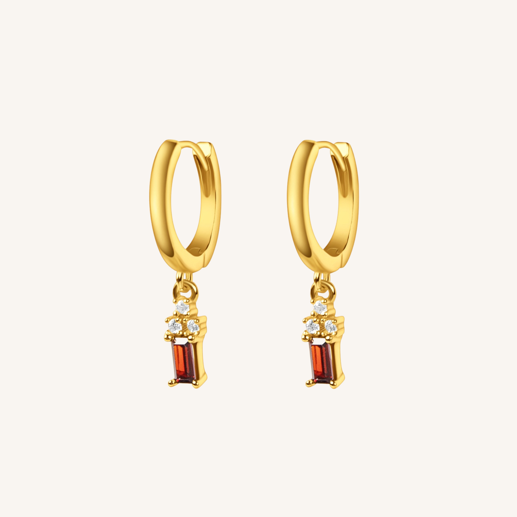 A pair of gold huggie hoop earrings with dangling rectangular red gemstone charms and small clear stones, shown on a light cream background.